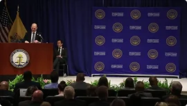 A man speaking at a podium with an audience and a backdrop displaying official seals and text.