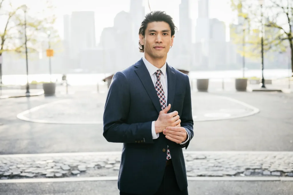 Professional man in navy suit and patterned tie standing outdoors with blurred city skyline background.